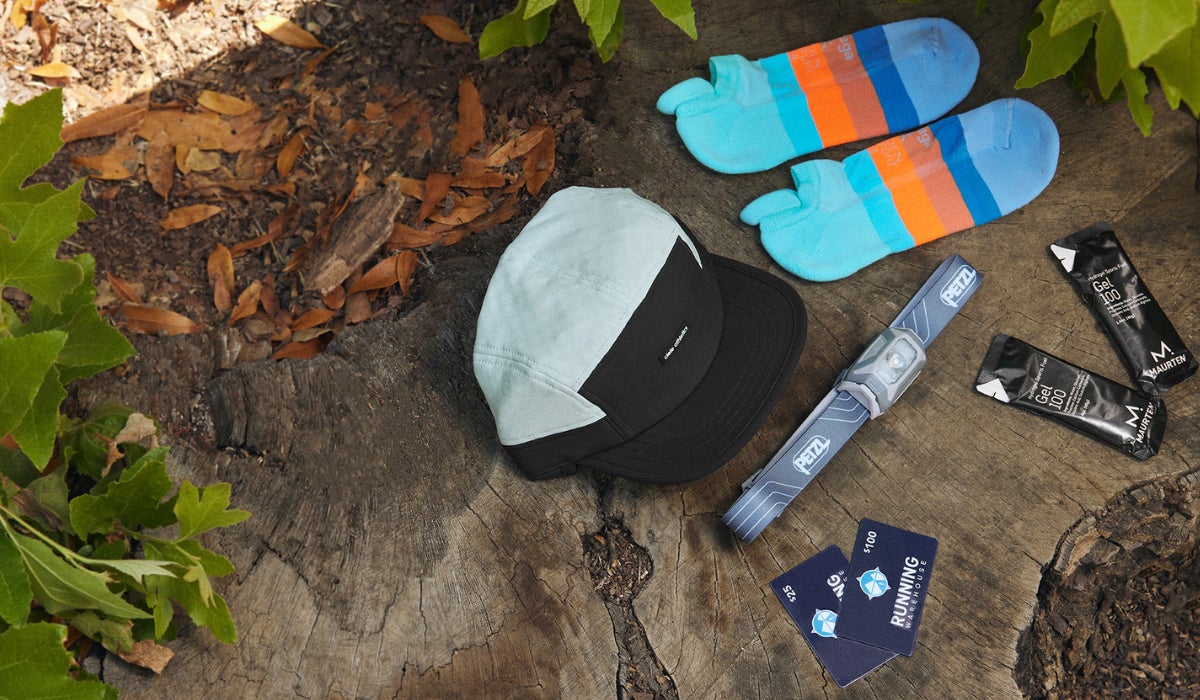 An array of running gifts on a tree stump, featuring a cap, blue gradient socks, a headlamp, energy gel packs, and race bibs. Surrounded by vibrant green leaves.