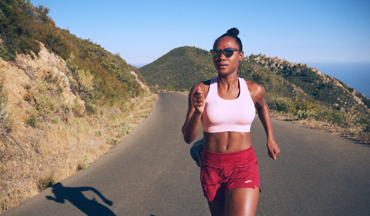 A woman runs confidently on a scenic mountain road wearing a pink sports bra and red shorts. The sun casts her shadow on the pavement, conveying focus and determination.
