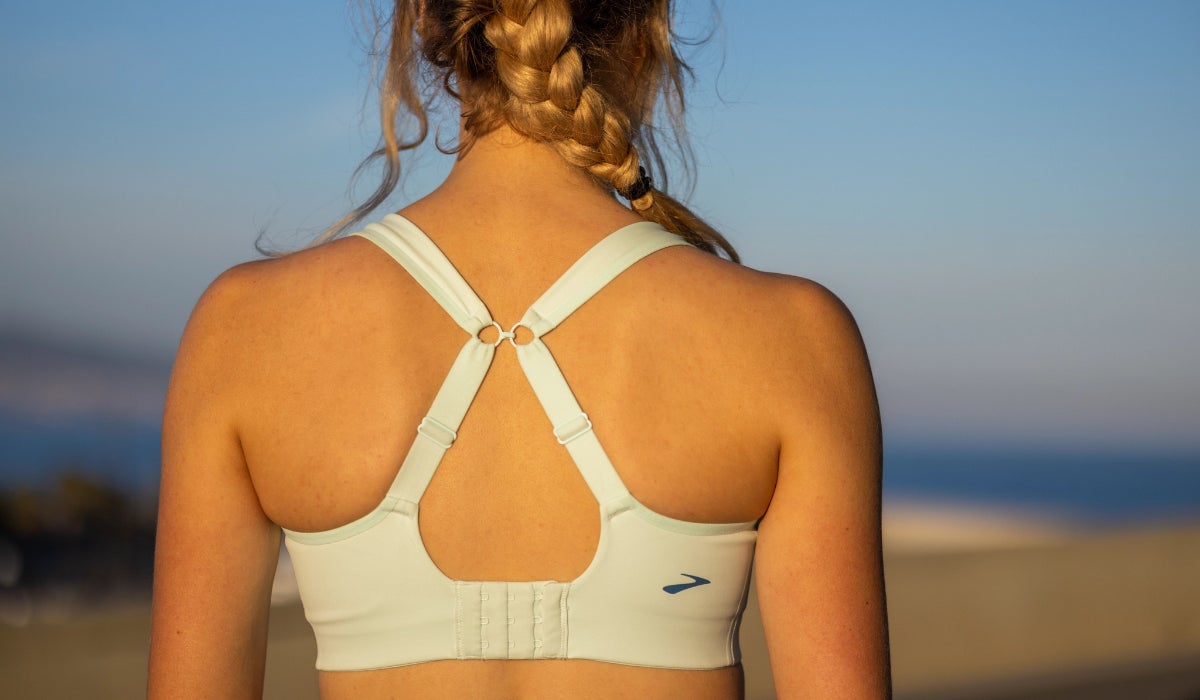A woman with braided hair is seen from behind, wearing a light gray sport bra. The scene is outdoors with a clear blue sky, conveying a sense of tranquility.