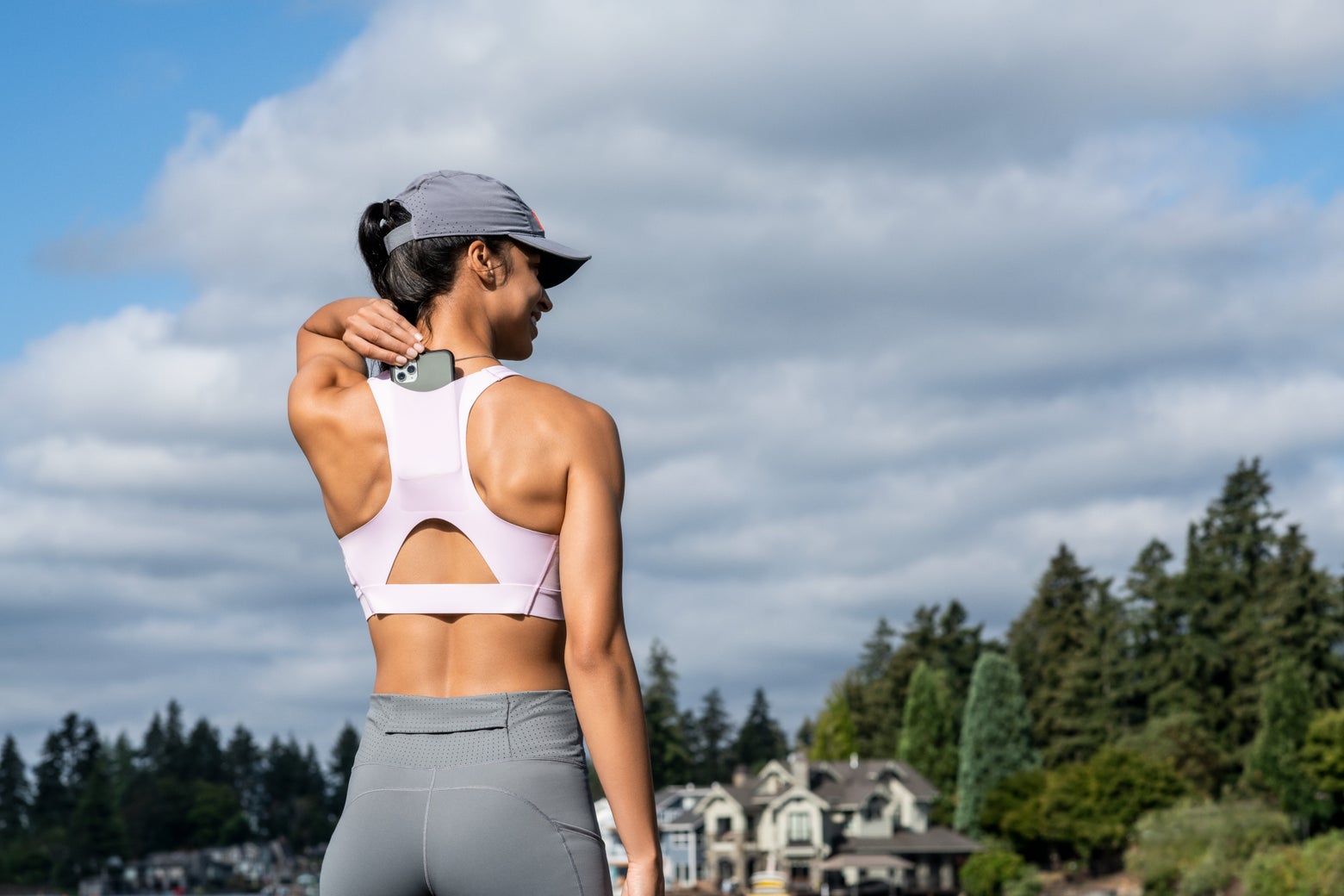 A woman in athletic wear and a cap stands outdoors, facing away, holding a massage device to her neck. The sky is cloudy, with trees and houses in the distance.