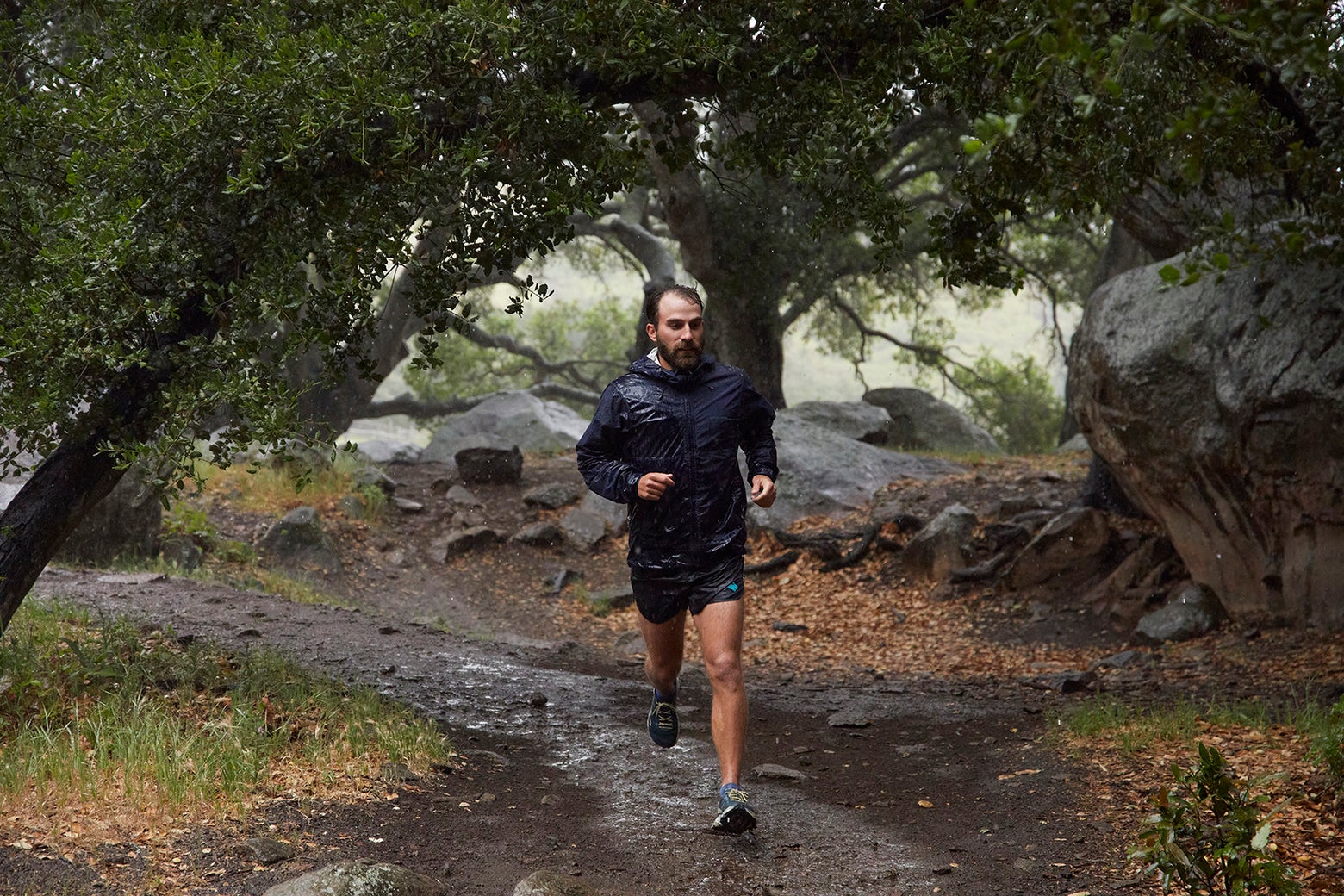 A bearded man jogs on a muddy forest trail during light rain, surrounded by lush green foliage and large rocks, conveying a sense of determination and serenity.