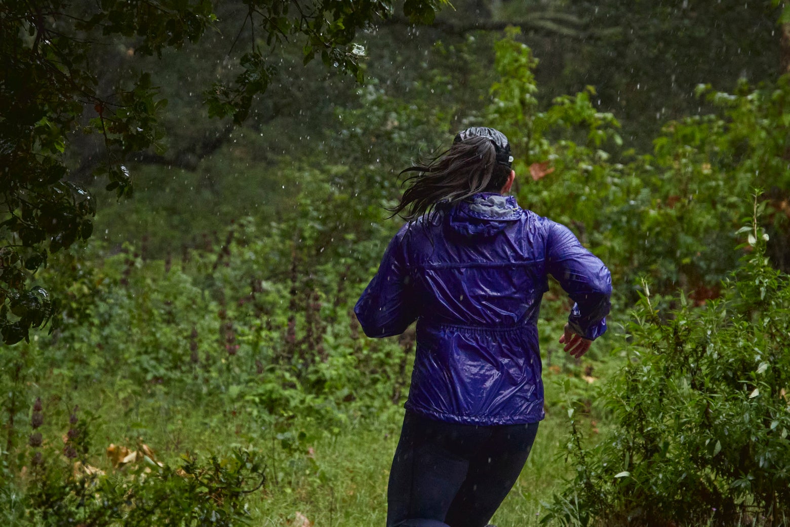 A person runs through a forest during rain, wearing a shiny blue jacket. The scene is lush and green, conveying a sense of adventure and determination.