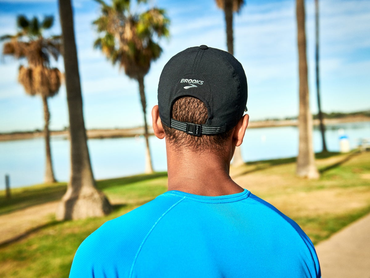A person in a blue shirt and black cap stands outdoors, facing a serene lake surrounded by palm trees under a bright blue sky. Peaceful and sunny.
