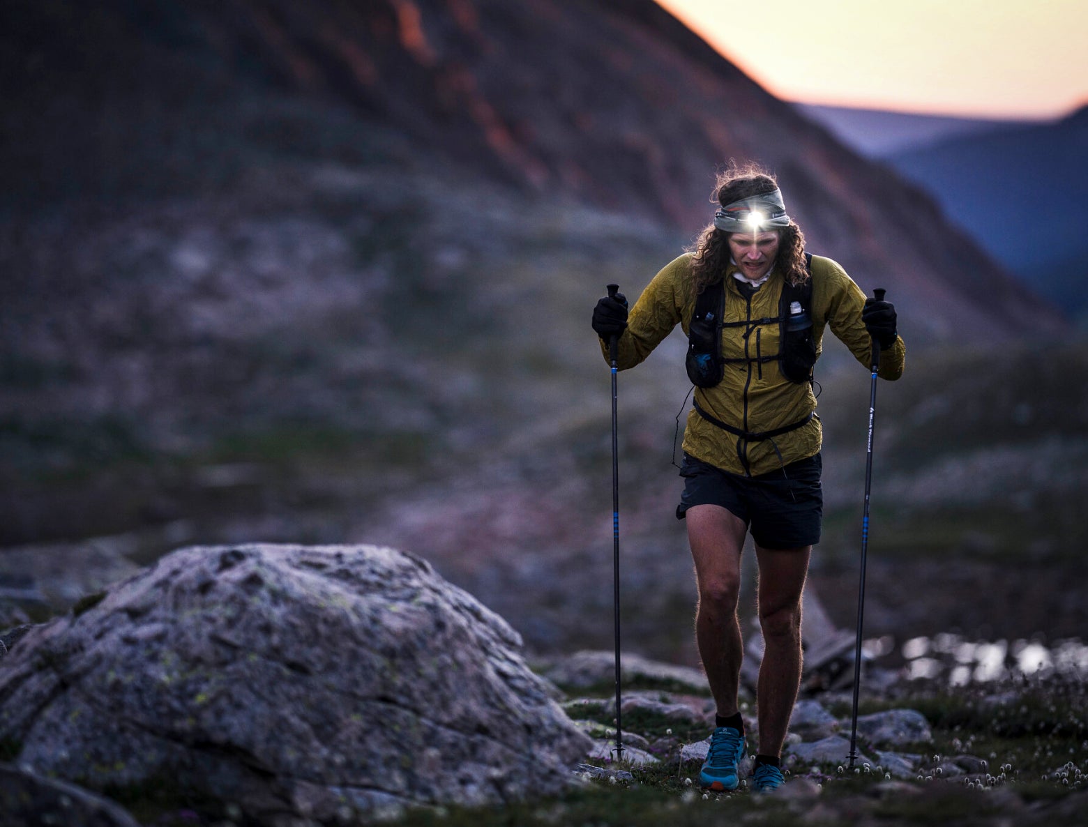 A person hikes uphill in a rocky landscape at sunrise, wearing a headlamp, yellow jacket, and backpack. The scene conveys determination and focus.