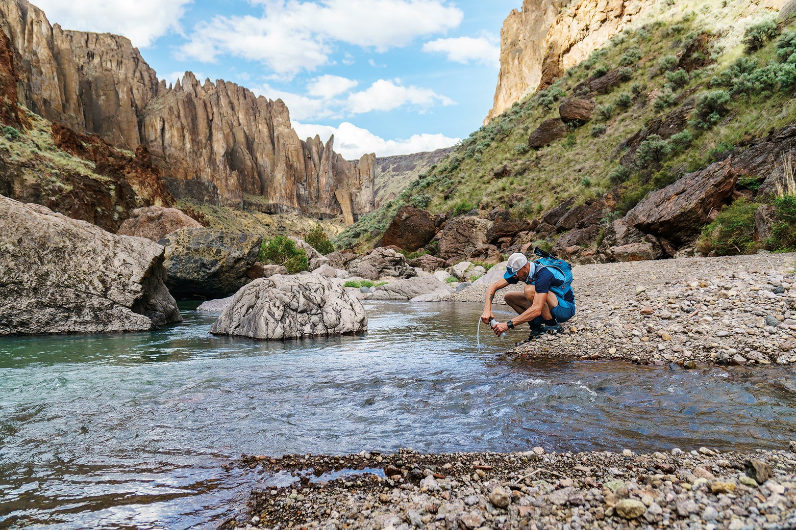 A hiker in blue outdoor gear kneels by a clear river in a rocky canyon, collecting water. The scene is bright and tranquil, with a blue sky above.