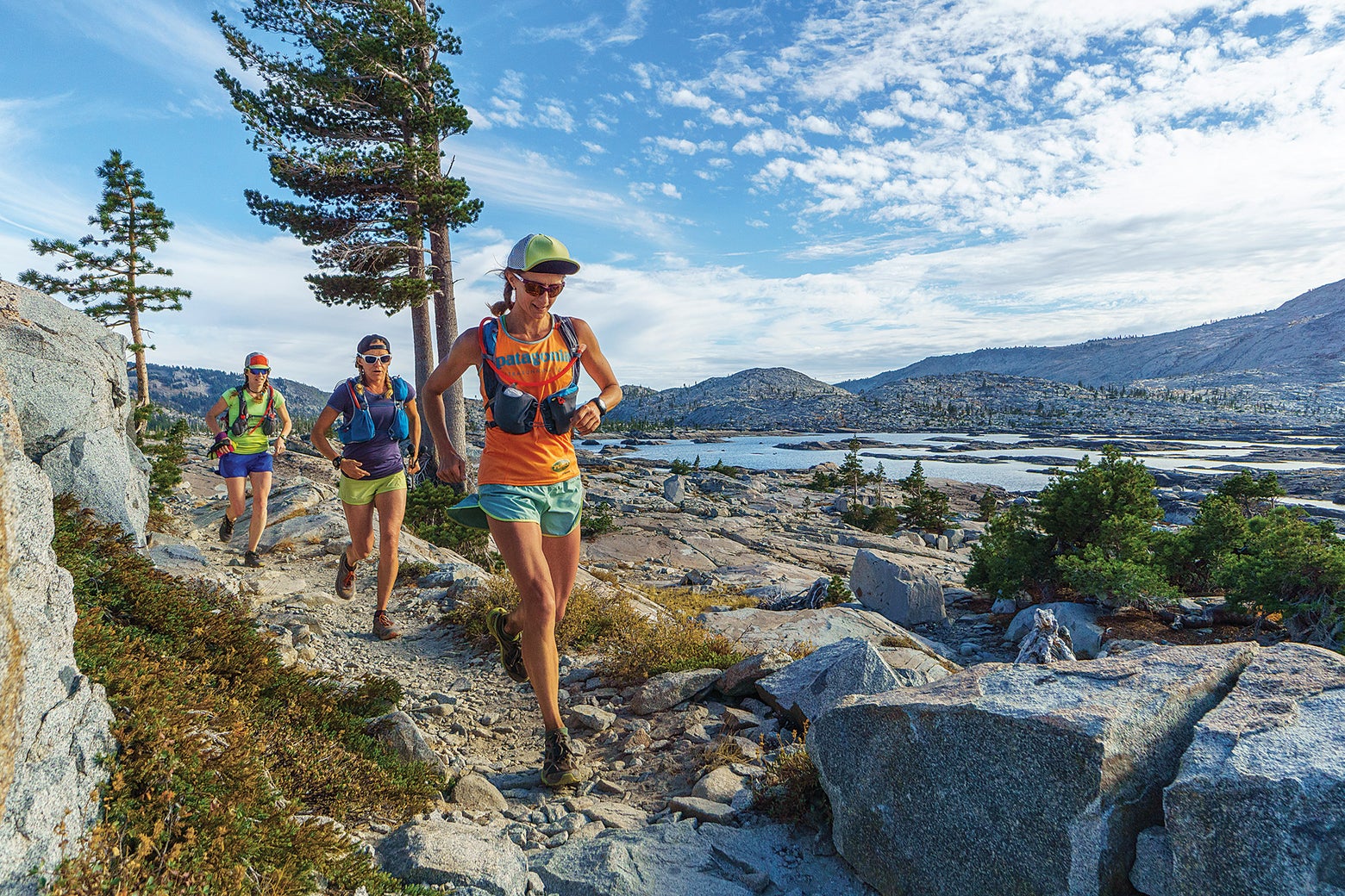 Three people trail running on a rocky mountain path beside a lake. They display a joyful, energetic mood under a bright blue sky.