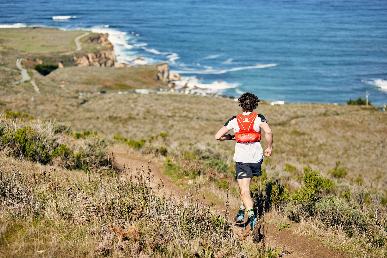 A person wearing a red vest runs on a coastal trail with ocean waves crashing. The scene is sunny and energetic, conveying freedom and adventure.