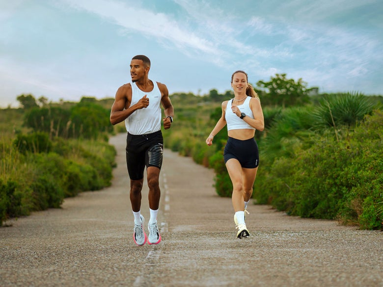 Two runners running on paved road surrounded by greenery, both wearing Kiprun apparel and Kipstorm shoes