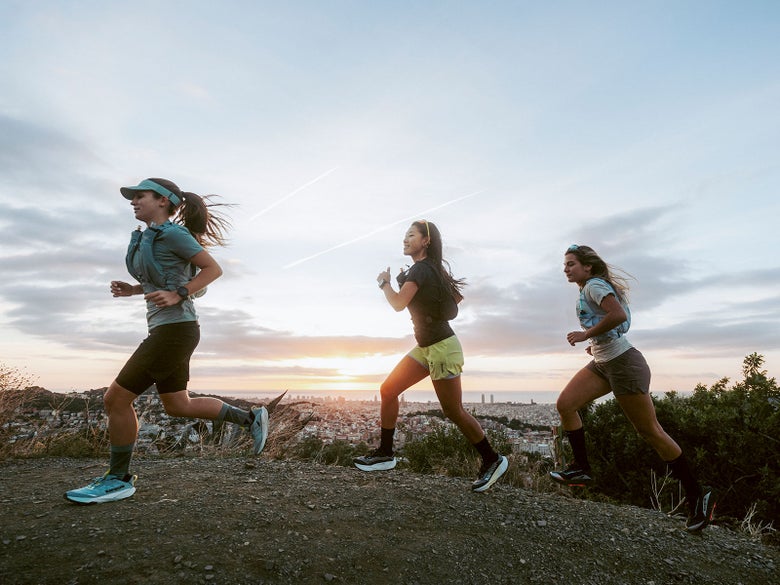 Three athletes running on a trail with a cityscape behind them. They are each wearing a different Kipsummit shoe. 