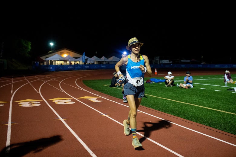 Magda Boulet finishing the Western States 100 race on a track at night, wearing a straw hat and a blue tank top. Spectators sit on the grass, illuminated by bright lights.
