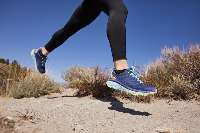 Runner in black leggings and blue HOKA Challenger trail shoes leaps over a sandy path, surrounded by desert shrubs under a clear blue sky, conveying energy and freedom