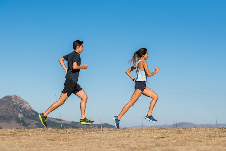 Image of a male and female runner running outside on a dirt path, there are clear blue skies and a green mountain/hill in the distance. 