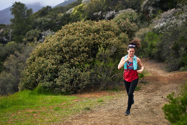 A woman jogs on a scenic dirt trail surrounded by lush green bushes and hills, wearing a red top and hydration pack while holding energy chews. The mood is energetic and peaceful.