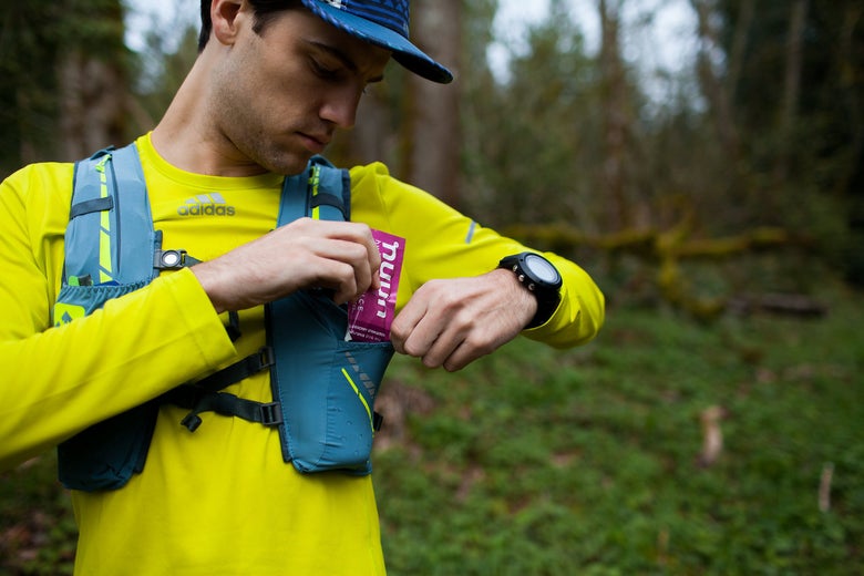 A man in a bright yellow shirt and blue cap retrieves a purple energy packet from a teal chest pouch. He stands in a lush forest, conveying a focused mood.