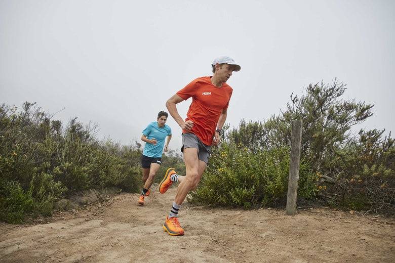 Hayden Hawks running on a trail in the Hoka Tecton shoes and a red shirt on a foggy path, surrounded by shrubs, conveying determination.