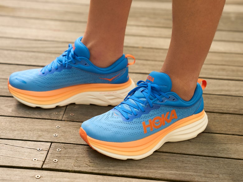 Close-up of a person wearing bright blue and orange Hoka running shoes, standing on a wooden deck. The scene conveys a sporty and energetic vibe.
