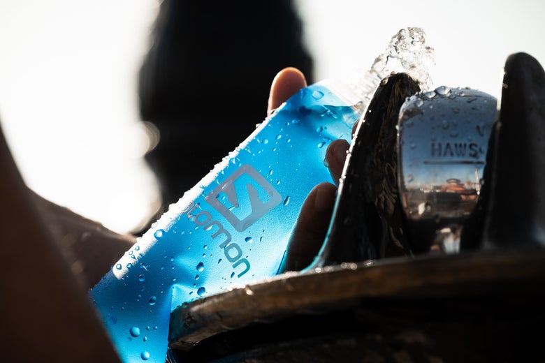 Hand holding a blue water bottle under a drinking fountain. Sunlight glints off the bottle as water splashes, conveying a refreshing, vibrant scene.