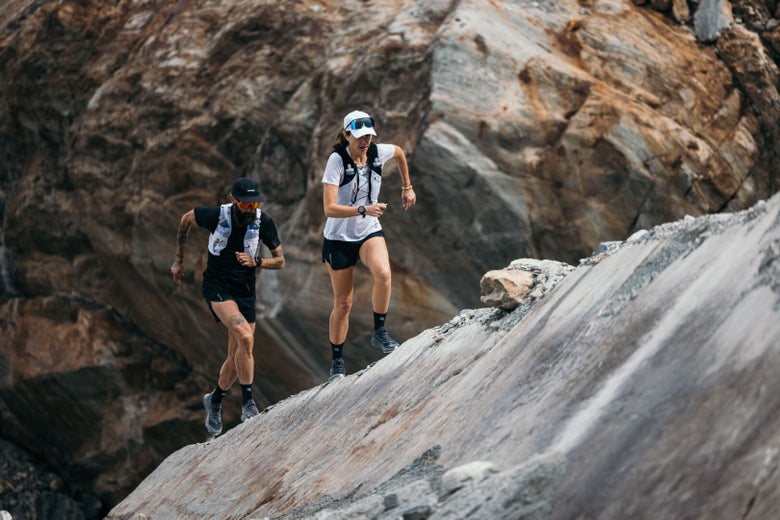 Two runners in athletic gear and backpacks ascend a rocky slope with determination, set against a backdrop of rugged cliffs and rocks.
