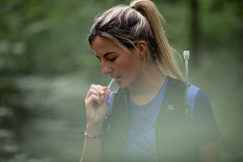 A woman in a blue striped shirt drinks from a hydration pack tube while hiking in a forest. Her expression is focused. Lush green trees surround her.