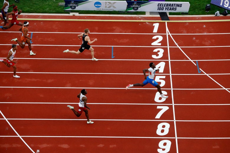 Overhead view of sprinters finishing a race on a red track. The runner in lane 5 crosses the finish line first, showing determination and speed.