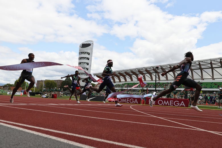 Runners sprint towards the finish line on a track under a partly cloudy sky. The first athlete breaks the tape, conveying intense competition and excitement.