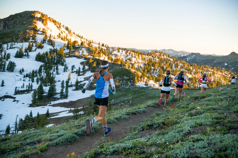 Trail runners traverse a scenic mountain path at sunrise. Snow patches and pine trees dot the landscape, with an atmosphere of adventure and determination.