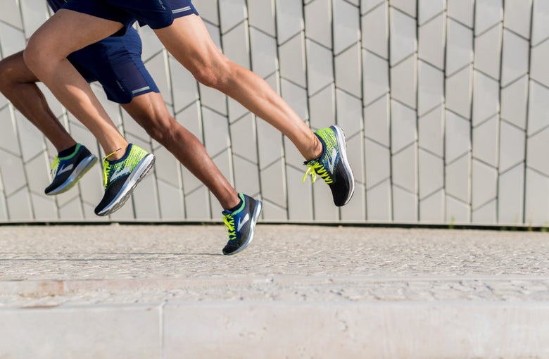 Two runners in motion, wearing blue shorts and neon-accented running shoes, on a textured pavement with a geometric-patterned wall behind them. Dynamic and energetic.