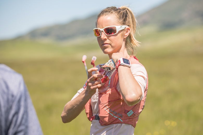 A woman wearing sporty sunglasses and a hydration vest stands in a grassy field under a clear sky, looking focused and ready for outdoor activity.