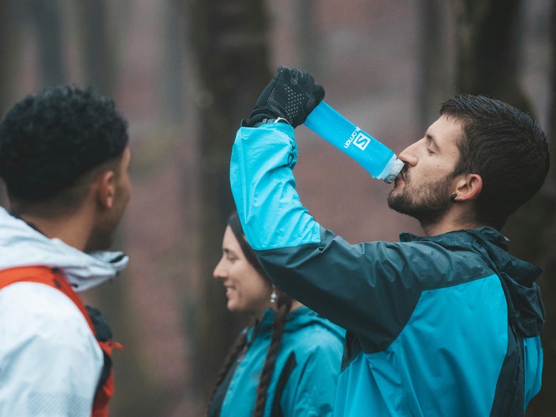 A group of three people in outdoor gear are in a forest. One person drinks from a blue water bottle, and two others smile in conversation. It's a relaxed atmosphere.