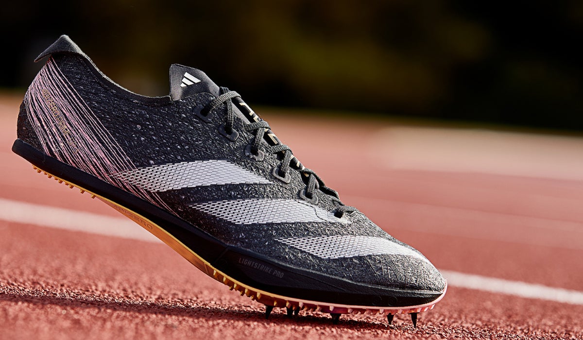 A sleek black Adidas athletic spike shoe with white stripes is displayed on a red running track. The shoe's dynamic design conveys speed and performance.