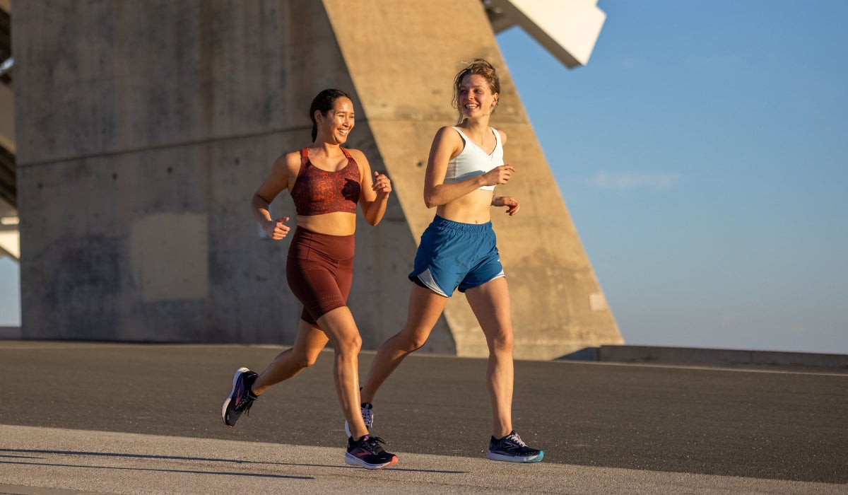 Two women jog on a sunny day under a modern concrete structure. One wears a maroon sports outfit, the other a white top and blue shorts. They are smiling.