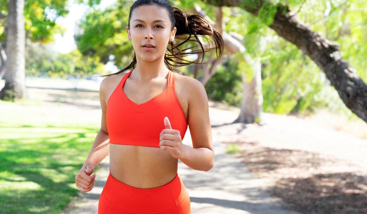 A woman jogs on a sunny, tree-lined path, wearing an orange sports bra and leggings. She appears focused and determined, embodying a sense of vitality.
