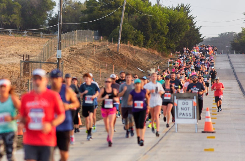 A large group of diverse runners participates in a road race, moving uphill on a wide path lined with traffic cones and a