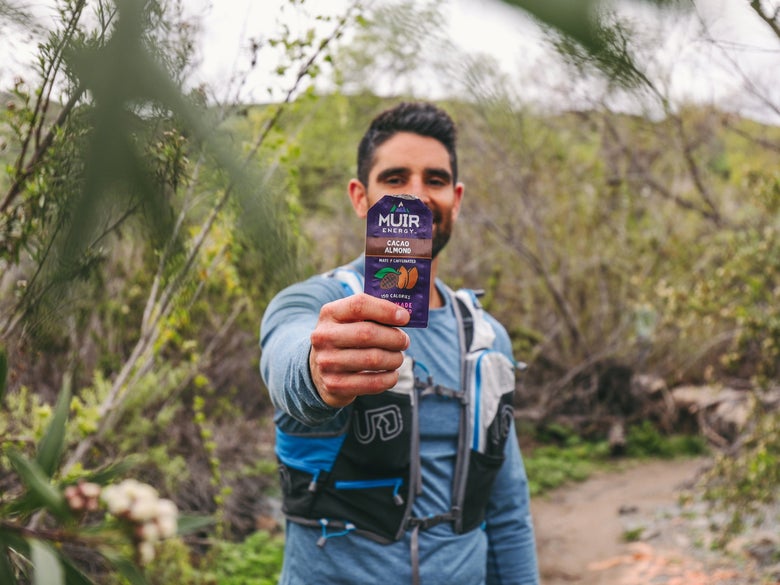 A man in hiking gear holds a Muir Energy cacao almond packet towards the camera. He's on a trail surrounded by lush greenery, conveying an adventurous vibe.