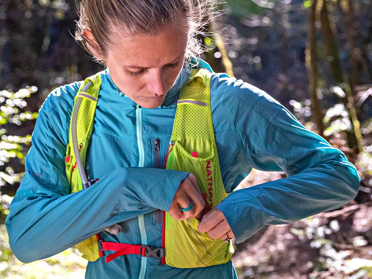 Woman in a blue jacket adjusts a yellow running vest in a sunlit forest. She appears focused as dappled light filters through the trees.