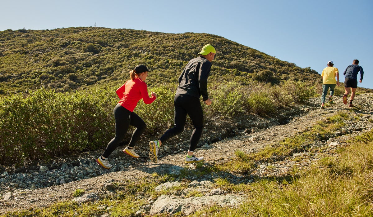 Four people trail running uphill on a rocky path against a backdrop of green hills and clear blue sky, conveying energy and determination.