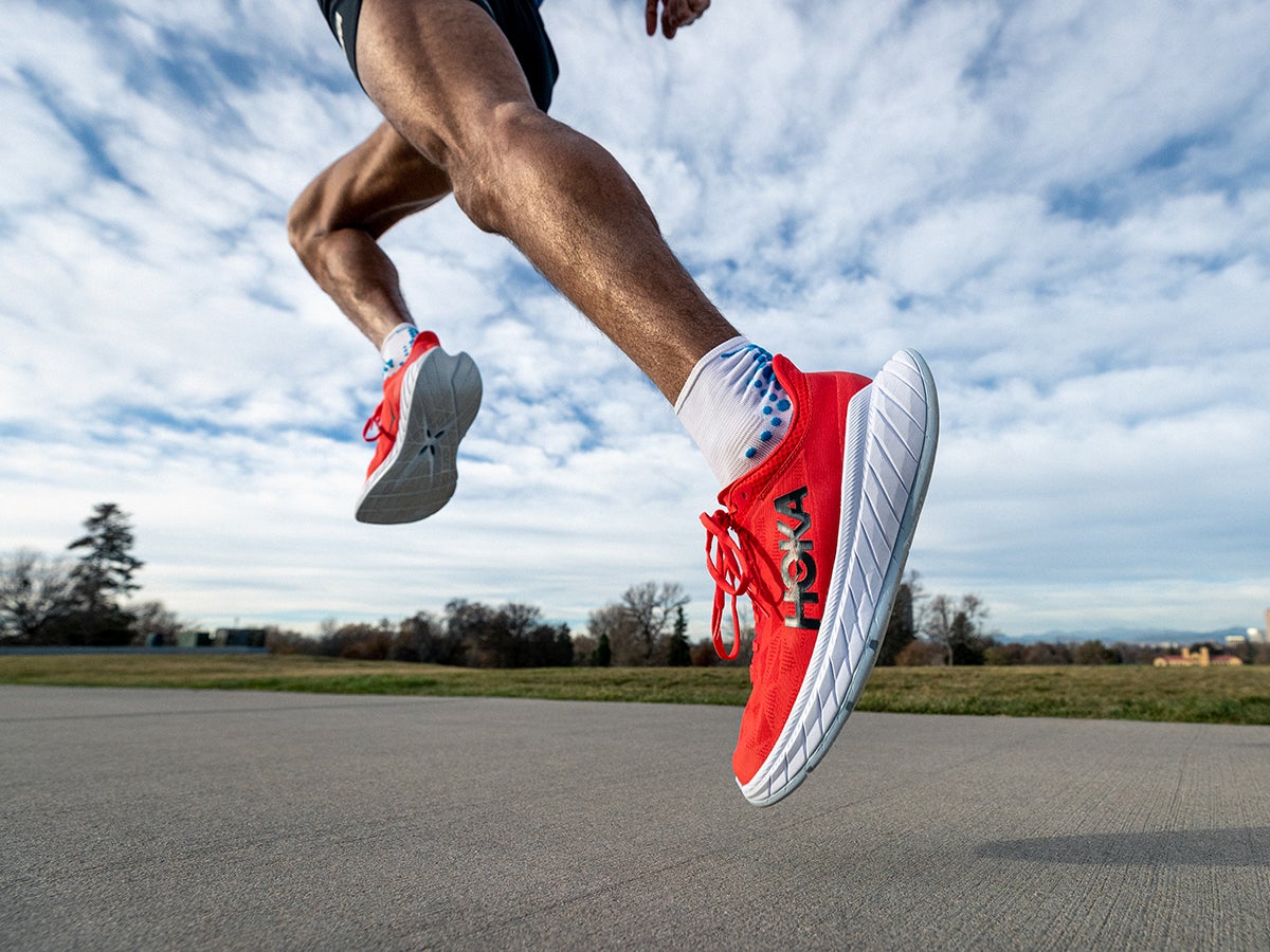 Runner in vibrant red HOKA sneakers strides on a concrete path under a partly cloudy sky, conveying energy and motion in a scenic outdoor setting