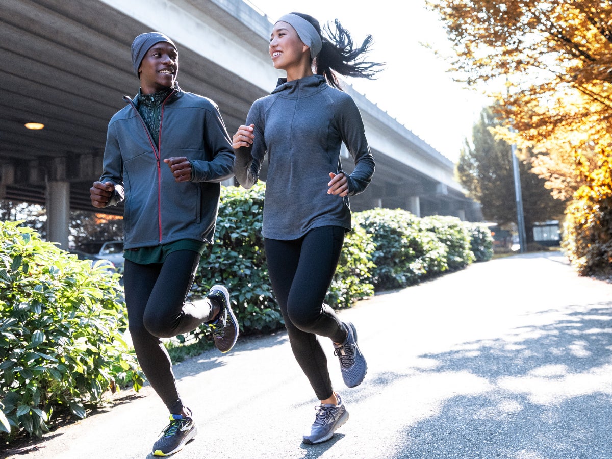 Two people jogging on a sunny path lined with green bushes and autumn trees, wearing athletic gear and smiling, conveying a cheerful mood.