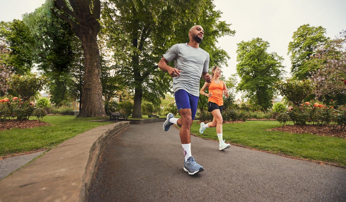 Two people jogging on a park path, surrounded by lush green trees and flowers. Both appear focused and energized under a partly cloudy sky.