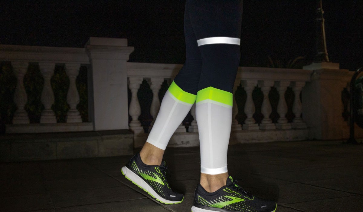 A person jogging at night wearing reflective leggings and running shoes. The bright reflectors and neon accents stand out against the dark background, ensuring visibility.