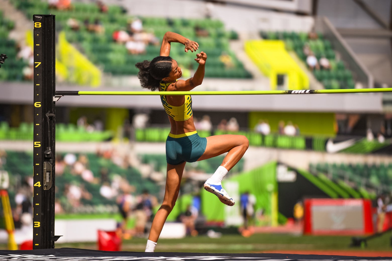 Athlete mid-air during a high jump, wearing a green and yellow uniform, against a vibrant stadium backdrop. The scene conveys determination and motion.
