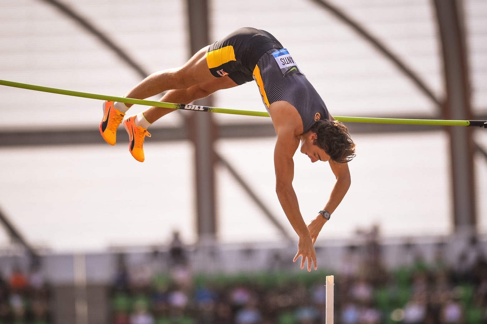 Athlete wearing a black and yellow uniform vaults over a high bar, displaying strength and concentration, with blurred spectators and structure in the background.
