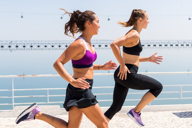 Two women jog energetically along a waterfront, wearing athletic gear. The background features calm water and a distant bridge under a clear sky.