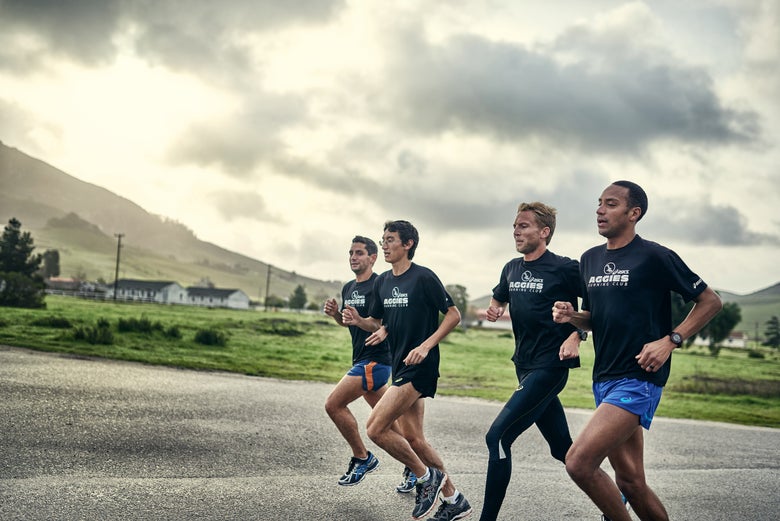 Four men jog together on a rural road, wearing matching sports attire. The sky is overcast and the background features rolling green hills.