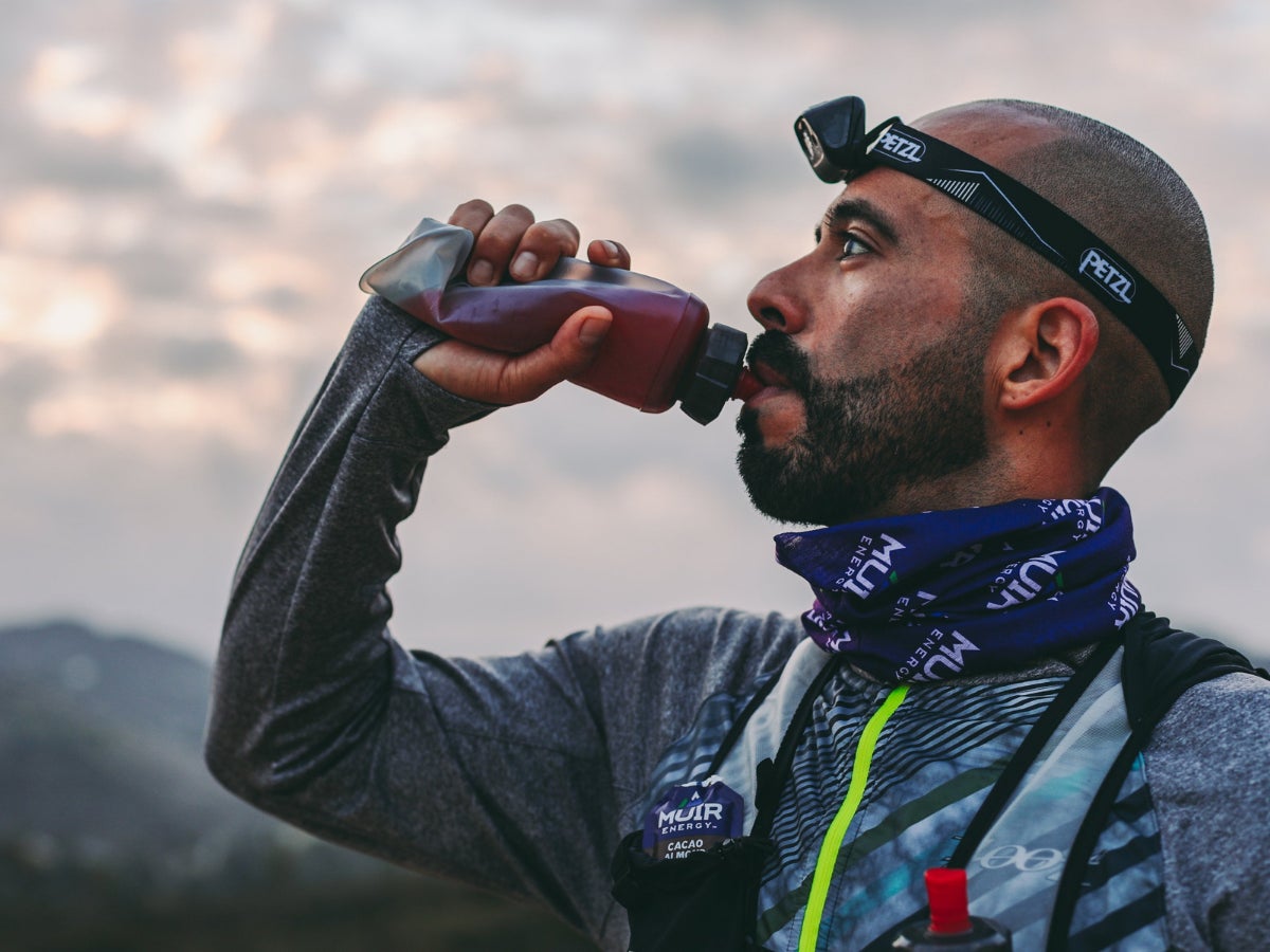 Man in outdoor gear and headlamp drinks from a bottle against a cloudy sky backdrop. He wears a purple neck gaiter, conveying focus and determination.