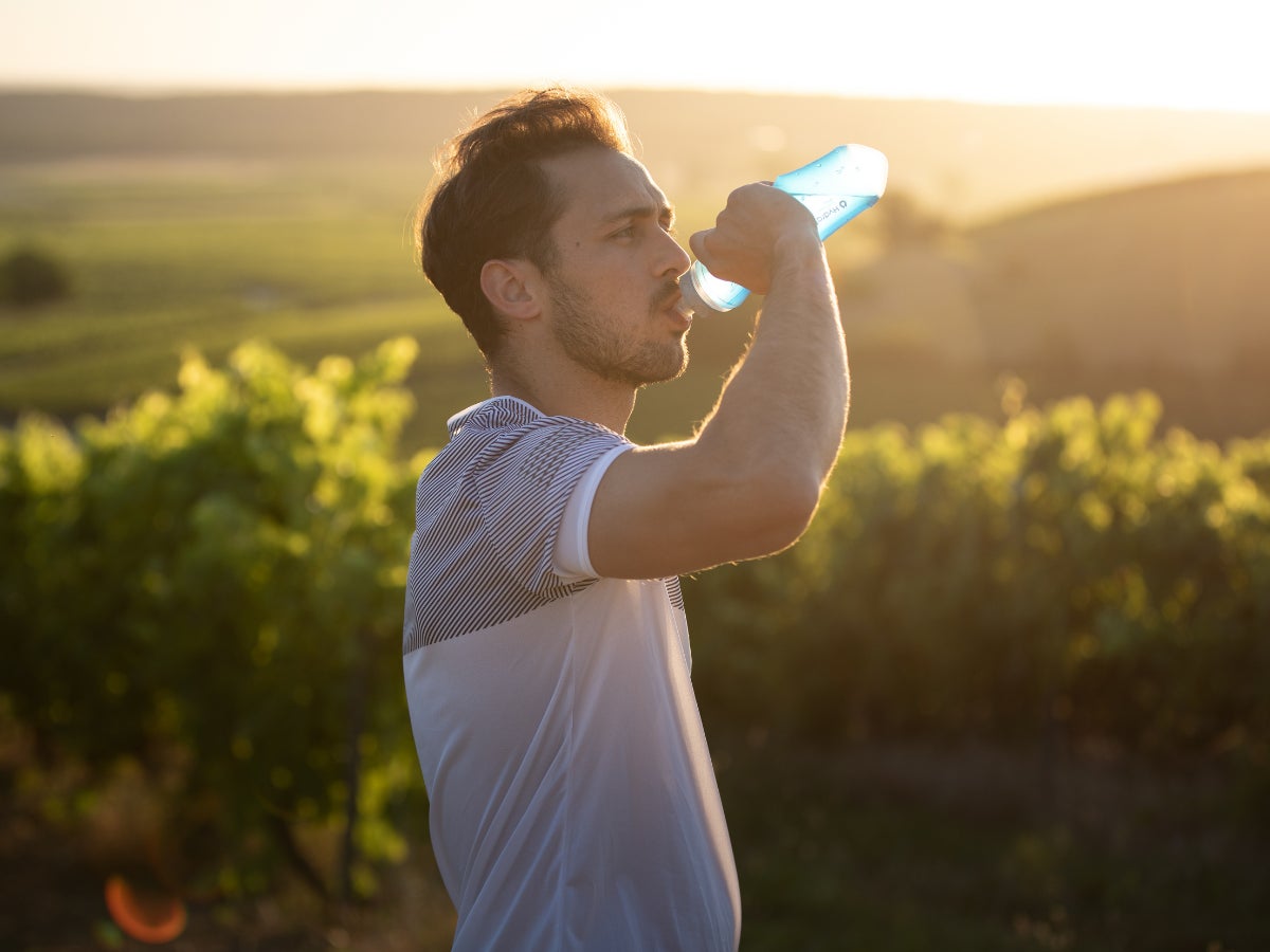 A man drinks from a soft flask bottle while standing in a sunlit vineyard. The golden hour light casts a warm glow, creating a serene and refreshing atmosphere.