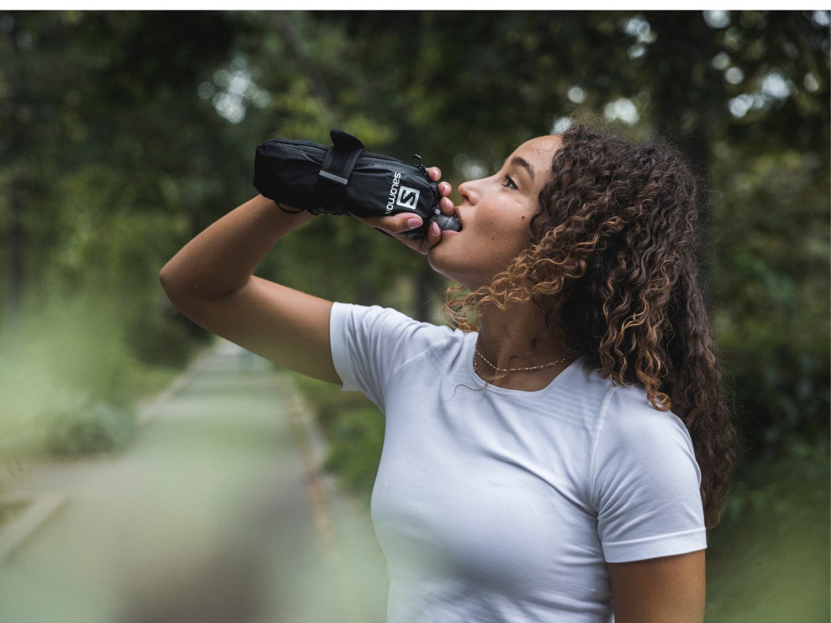 Woman in white shirt drinks water from a black bottle with a glove, standing outdoors. Green, blurred background conveys a refreshing, energetic tone.