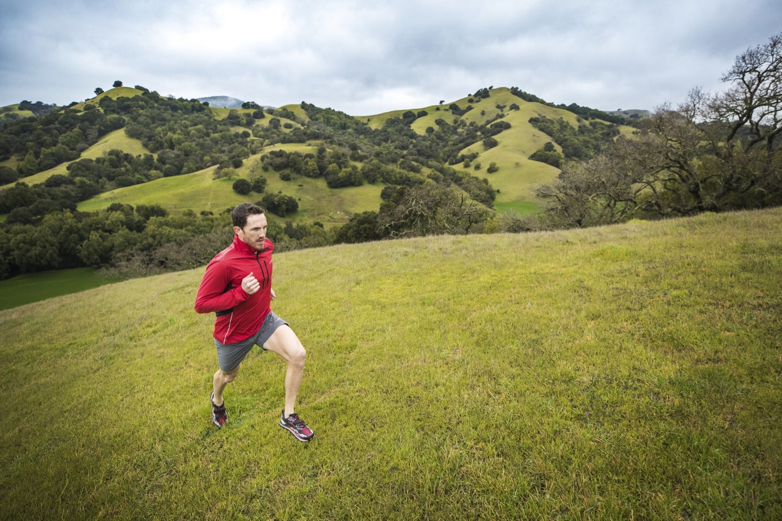 man running outside in a red jacket and gray shorts against the backdrop of green rolling hills