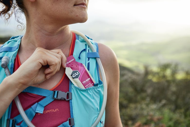 A woman in a turquoise hydration vest holds a GU energy gel. She's outdoors with a blurred green landscape behind, suggesting an active, adventurous mood.