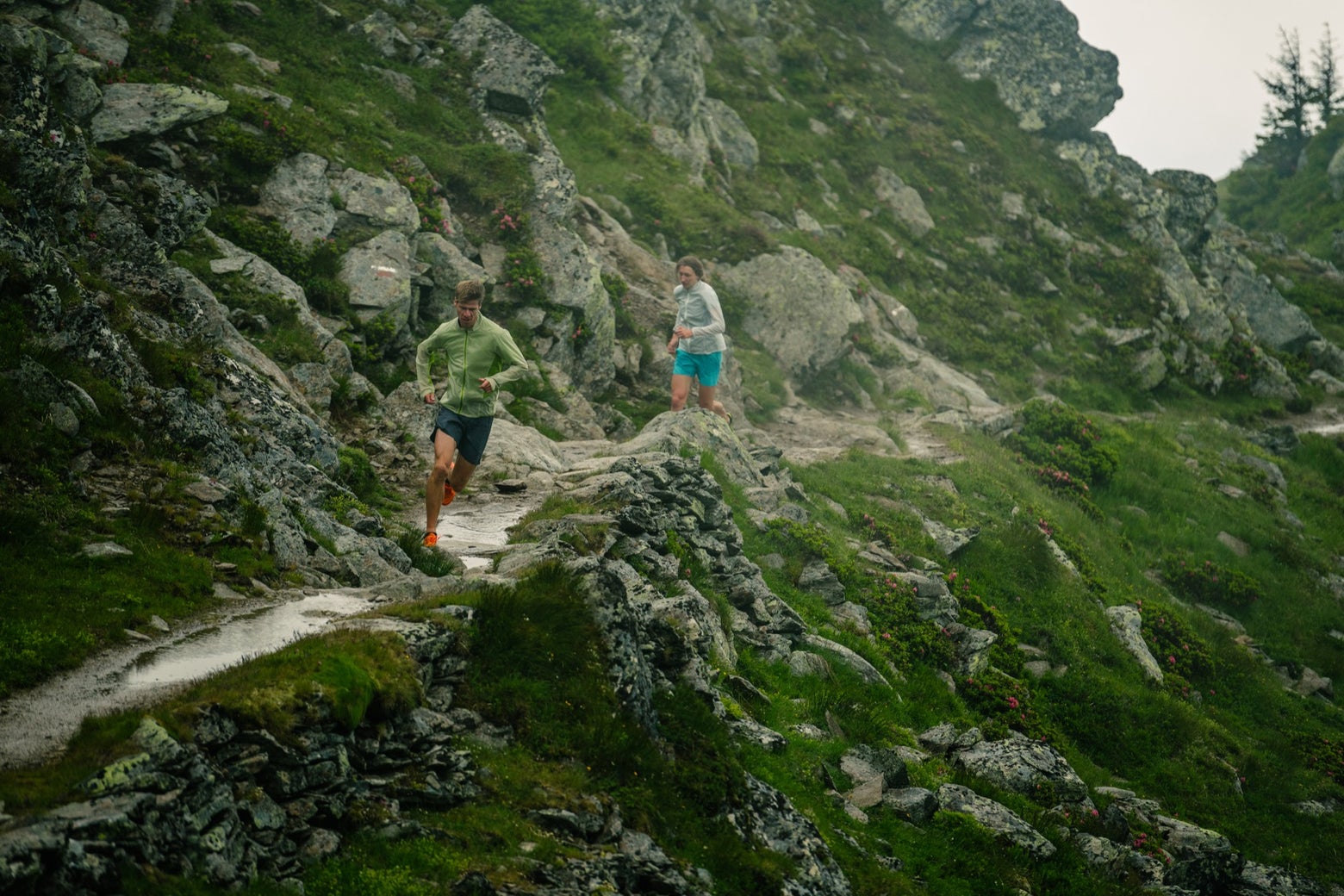 Two runners, male and female, running downhill on a rocky mossy trail with puddles